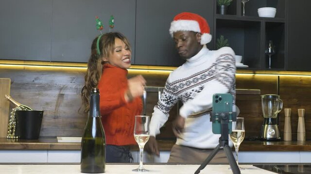 Funny african american couple in sweaters dancing in the kitchen in front of smartphone by video link, christmas party at home and virtual reality