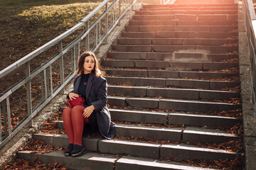 a woman in a dark green coat and red tights with a red handbag is sitting on the stairs in the park 