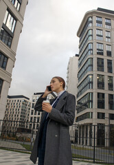 An office worker drinks coffee and talks on the phone on background of skyscrapers. An office worker went outside to buy coffee and talk on the phone. Young woman on the background of high-rise office