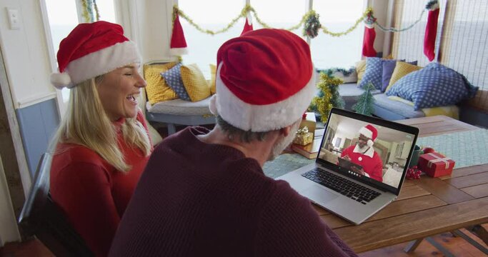 Smiling caucasian couple with santa hats using laptop for christmas video call with santa on screen