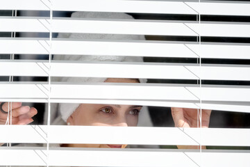 Middle aged woman with towel turban on her head pulls up the jalousie and looking through the window
