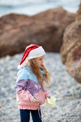 Little pretty girl in a santa claus hat on the seashore, a child walks in a jacket