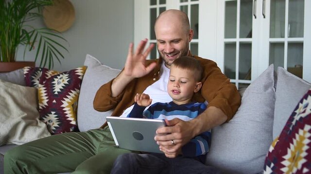 Father and son at videocall. Happy little boy with bearded man wave hands to tablet camera spbd communicating with relatives sitting on sofa in living room