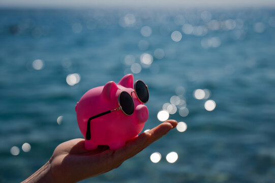 A woman holds a piggy bank in sunglasses on the background of the sea. Budget vacation.