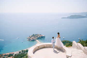 Fototapeta na wymiar The bride stands on the parapet of the observation deck overlooking the island of Sveti Stefan, the groom holds her hand