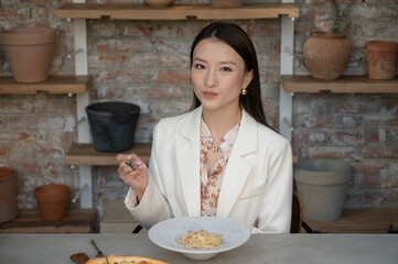 Portrait of stunning Chinese Asian woman in restaurant eating pasta and holding fork in her hand. Businesswoman in blouse enjoying lunch or dinner, old style industrial design cafe with clay pots