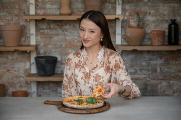 Portrait of stunning Chinese Asian woman in restaurant eating pizza and holding one slice in her hand. Businesswoman in blouse enjoying lunch or dinner, old style industrial design cafe with clay pots