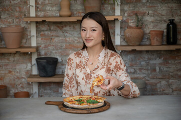Portrait of stunning Chinese Asian woman in restaurant eating pizza and holding one slice in her hand. Businesswoman in blouse enjoying lunch or dinner, old style industrial design cafe with clay pots