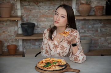 Portrait of stunning Chinese Asian woman in restaurant eating pizza and holding one slice in her hand. Businesswoman in blouse enjoying lunch or dinner, old style industrial design cafe with clay pots