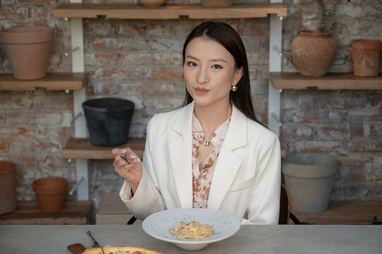 Portrait Of Stunning Chinese Asian Woman In Restaurant Eating Pasta And Holding Fork In Her Hand. Businesswoman In Blouse Enjoying Lunch Or Dinner, Old Style Industrial Design Cafe With Clay Pots