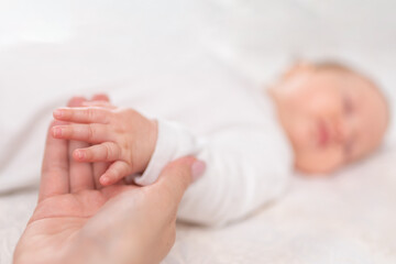 Young woman holding small hand of her sleeping baby daughter in bedroom