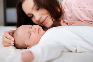 Fototapeta na wymiar Young woman lovingly looks at her sleeping newborn daughter in the bedroom