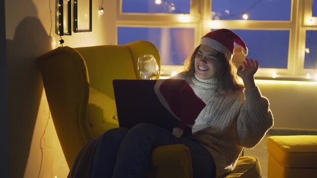 Cheerfull woman with curly hair making a toast with champagne glass during video call with friends or family, Portrait female in festive hat looking into laptop display, celebrating christmas online