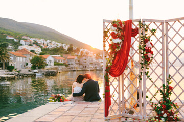 Newlyweds sit hugging each other on the pier against the backdrop of water, mountains and old houses. In the foreground there is an openwork screen decorated with flowers and a red curtain.