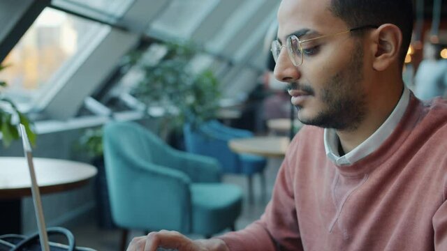 Slow motion tilt-down portrait of ambitious Middle Eastern guy working with laptop in cafe typing sitting at table alone. People and freelance job concept.