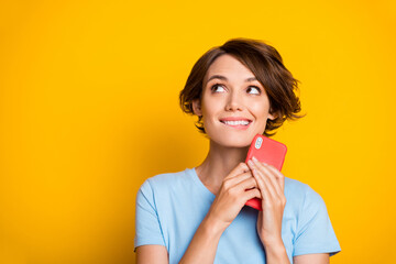 Photo of young girl happy positive smile dreamy look empty space bite lips hold cellphone isolated over yellow color background
