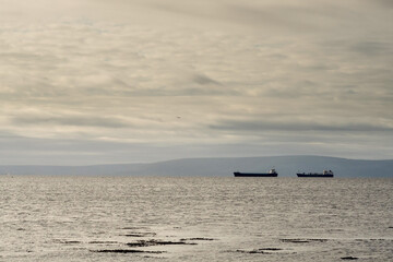 Silhouette of cargo ships in Galway bay waiting for high tide. International trade and business concept. Light pastel color. Burren mountains in the background. Selective focus. Transportation trade.