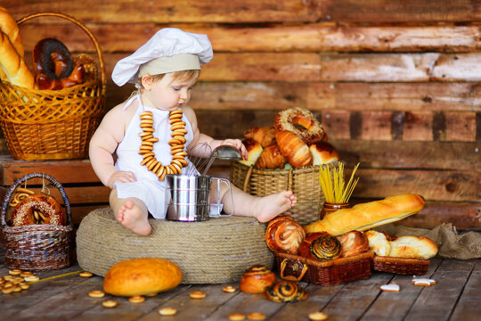 a baby boy in a chef's hat and apron with a bunch of bagels around his neck sits against the background of bakery products in a bread shop.