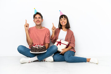 Young mixed race couple celebrating a birthday sitting on the floor isolated on white background showing and lifting a finger in sign of the best