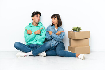 Young couple making a move while picking up a box full of things sitting on the floor isolated on white background pointing to the laterals having doubts