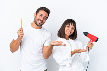 Young caucasian couple holding a hairdryer and toothbrush isolated on white background pointing back and presenting a product