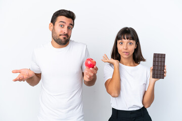 Young couple holding chocolate and apple isolated on white background having doubts while raising hands and shoulders