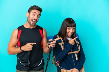 Young mountaineer couple with a big backpack isolated on blue background pointing finger to the side in lateral position
