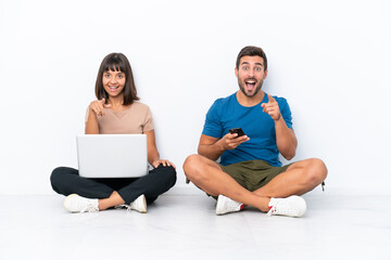 Young couple sitting on the floor holding pc and mobile phone isolated on white background points finger at you with a confident expression