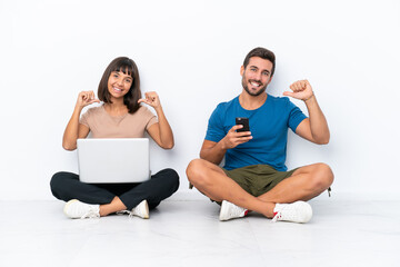 Fototapeta na wymiar Young couple sitting on the floor holding pc and mobile phone isolated on white background proud and self-satisfied in love yourself concept
