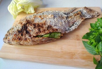 Authentic thai grilled fish (nile tilapia) on a wooden cutting board, garnished with coriander, basil and Chinese cabbage.