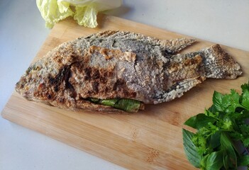 Authentic thai grilled fish (nile tilapia) on a wooden cutting board, garnished with coriander, basil and Chinese cabbage.