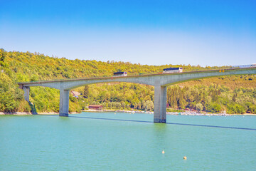 Buses ride on a concrete bridge over a lake with turquoise water against the backdrop of trees.