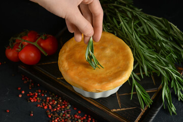 Woman decorating tasty vegetable pot pie with rosemary on black background
