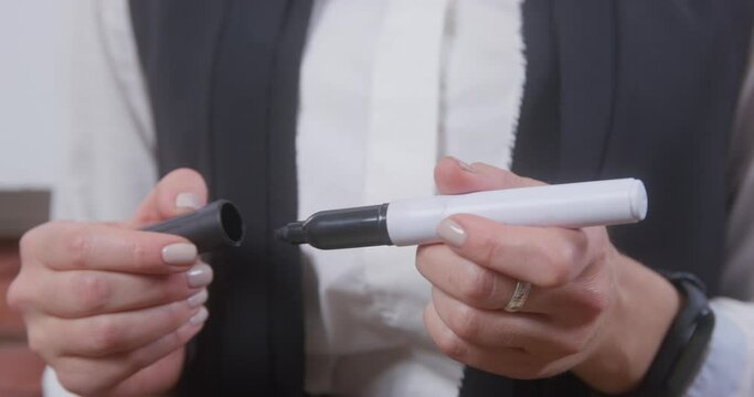 School teacher in white blouse with neat manicure stands IN classroom and opens-closes black permanent marker extreme closeup.