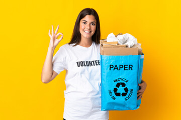 Young brazilian girl holding a recycling bag full of paper to recycle isolated on yellow background showing ok sign with fingers