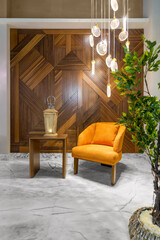 Modern orange armchair, illuminated wooden lantern on small wooden table, and contemporary tall glass chandelier, in a hall with decorated wood cladding wall, and white marble floor