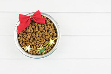 Metal bowl with dry pet food and Christmas decorations on light boards. Christmas pet supplies concept, New year and Christmas celebration. Top view, space to insert text.
