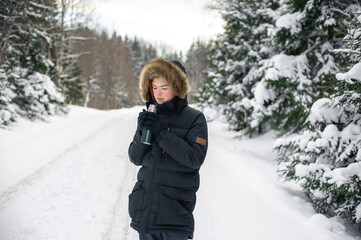 Fototapeta na wymiar Teenage boy walks through a snowy forest on a cold winter day with a hot drink in a thermos. Healthy lifestyle. Useful activity.