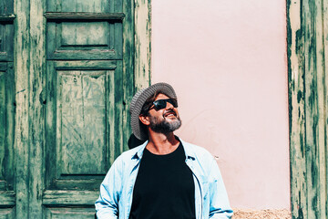 Hipster tourist smile at the sun standing against an old wall with green door and window. Happy adult mature man people with hat and sunglasses enjoying the lifestyle