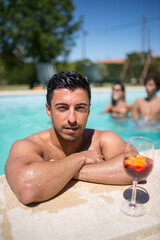 Sporty man drinking bright cocktail in swimming pool. Man with dark hair and glass with bright beverage, looking at camera. Leisure, friendship, party concept