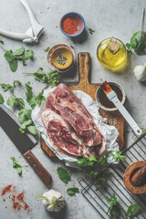Raw meat pieces on wooden cutting board with oil, spices, marinade, herbs and cooking utensils on grey kitchen table. Cooking and grill preparation at home with flavorful homemade marinade. Top view.