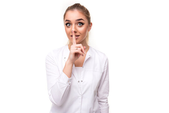 Photo of a beautiful pretty cute attractive positive young blonde woman with a ponytail and makeup in a white medical coat of a nurse showing shhh and secret gesture with one finger isolated on a
