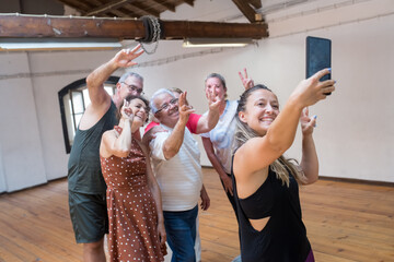 Happy young Caucasian dance teacher taking selfie with seniors.Hilarious instructor with long fair hair taking picture of her group of senior dancers in studio. Dance, hobby, healthy lifestyle concept