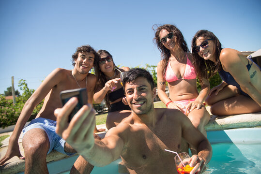 Happy friends taking selfie at poolside. Women and men in swimsuits resting in swimming pool, taking pictures with camera. Leisure, friendship, party concept