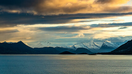 Fototapeta na wymiar Wilde, schneebedeckte Berge in der Umgebung von Ørnes, Norwegen, im orangen Morgenlicht bei Sturm mit Wolken. Schneefahnen wehen von den Bergspitzen herunter. Kreuzfahrt mit dem Postschiff 