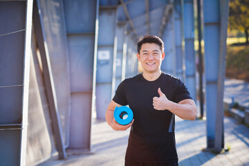 Portrait of asian athlete with mat for him near stadium looking at camera and smiling holding thumbs up yoga instructor