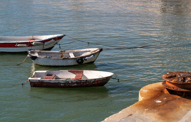 View of three old flaked wooden boats moored to a pier on the right with a part of a rusty bollard on the concrete surface in a defocused foreground, warm sunny day, Cascais, Portugal