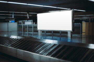 View of a baggage claim area in a hall of a modern airport arrival zone with luggage conveyor belt, with a selective focus on a template of a blank advertising poster or an information billboard