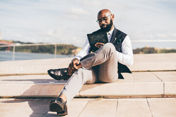 A handsome bald bearded black man entrepreneur in eyeglasses and a formal suit with a vest, is sitting outdoors and interacting with a futuristic tablet pc gadget with infographics on its screens