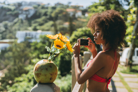 A side profile of a gorgeous young African-American female in a red bikini, with fluffy afro hair, standing in a resort area near a tropical beach and shooting panoramic images on her smartphone